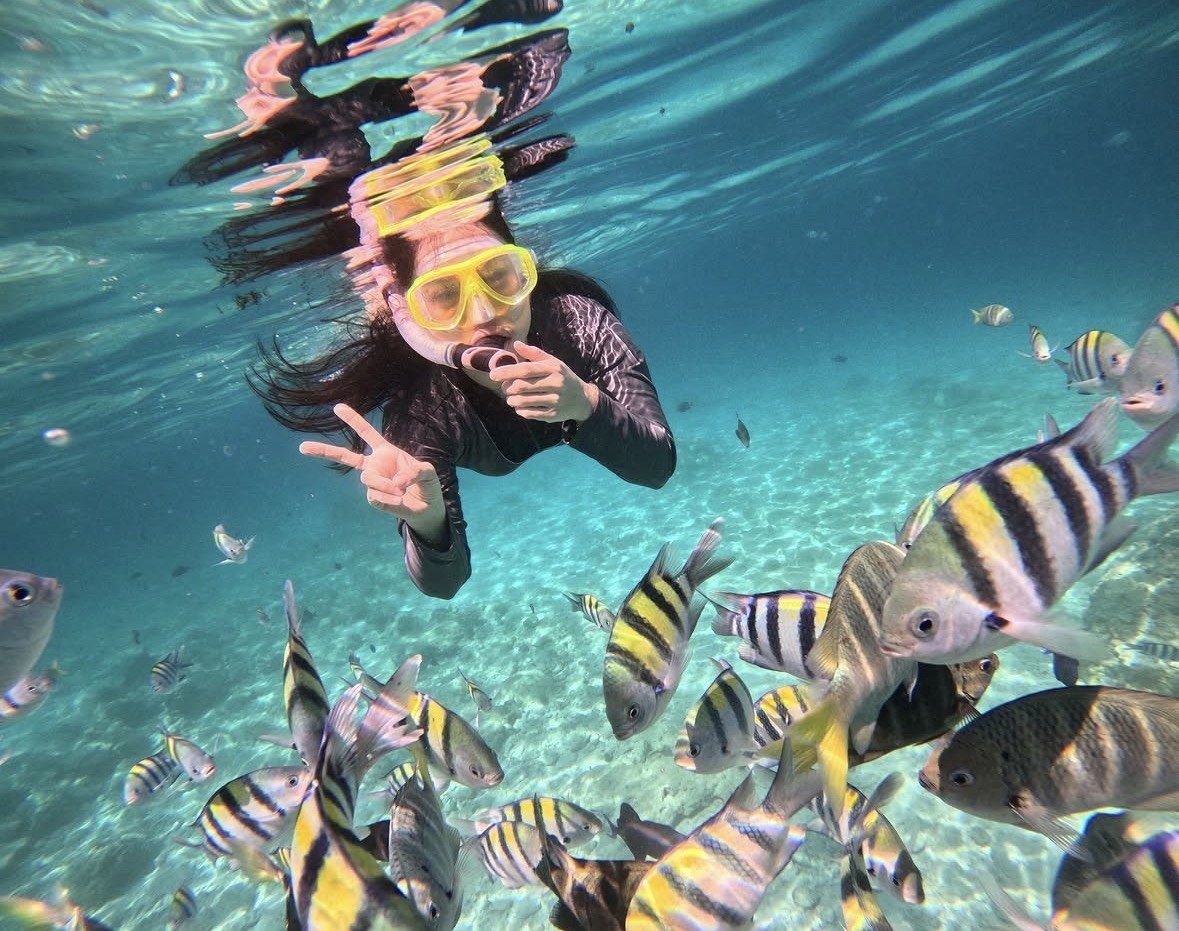Woman in black wetsuit flashing a peace sign among sergeant major fish